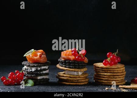 Stapel von schwarzen Vollkorn Holzkohle und traditionellen Cracker mit Räucherlachs, Frischkäse, grüner Salat und rote Johannisbeere Beeren über schwarzen Stein bac Stockfoto