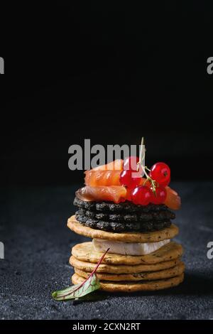 Stapel von schwarzen Vollkorn Holzkohle und traditionellen Cracker mit Räucherlachs, Frischkäse, grüner Salat und rote Johannisbeere Beeren über schwarzen Stein bac Stockfoto