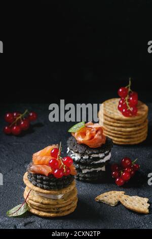 Stapel von schwarzen Vollkorn Holzkohle und traditionellen Cracker mit Räucherlachs, Frischkäse, grüner Salat und rote Johannisbeere Beeren über schwarzen Stein bac Stockfoto