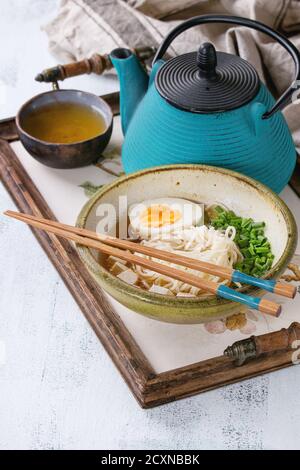 Schüssel mit asiatischer Suppe mit Nudeln, Ei, Frühlingszwiebeln und Tofu-Käse, serviert mit Essstäbchen, Teekanne und Tasse grünem Tee auf Holztablett mit textil Stockfoto