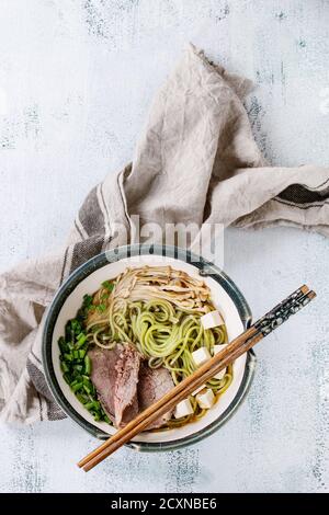 Schüssel asiatische Suppe mit grünem Tee Soba Nudeln, Pilze, Rindfleisch, Frühlingszwiebeln und Tofu-Käse, serviert mit Essstäbchen und Textil über weißem Woo Stockfoto