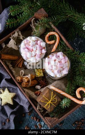 Zwei Gläser Café Latte mit rosa Marshmallow stehend in Holzkiste mit Dekor, Bonbons, Gewürze, Kaffee und Tanne Weihnachtsbaum über dunkle Textur ba Stockfoto