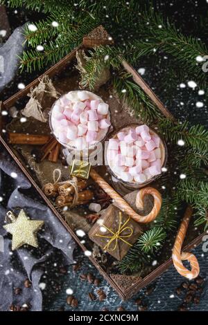 Zwei Gläser Café Latte mit rosa Marshmallow stehend in Holzkiste mit Dekor, Bonbons, Gewürze, Kaffee und Tanne Weihnachtsbaum über dunkle Textur ba Stockfoto
