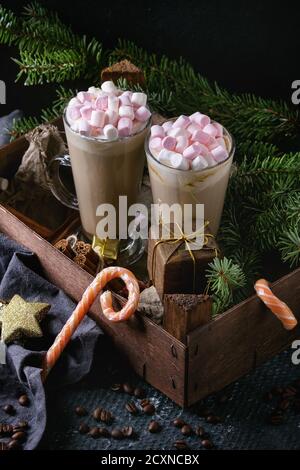 Zwei Gläser Café Latte mit rosa Marshmallow stehen in Holzkiste mit Weihnachtsdekor, Süßigkeiten, Gewürzen, Kaffeebohnen und Tannenbaum über dunklem Text Stockfoto