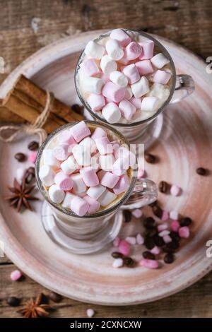 Zwei Gläser Café Latte mit rosa Marshmallow auf Keramikplatte mit Gewürzen, Kaffeebohnen und rosa Zucker über Holz Textur Hintergrund stehen. Nach oben Stockfoto