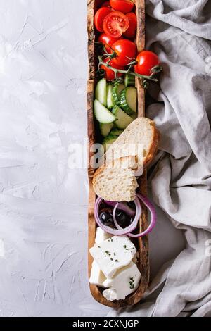 Zutaten für traditionellen griechischen Salat. Kirschtomaten, Gurken in Scheiben geschnitten, rote Zwiebeln, schwarze Oliven, Feta-Käse in Olivenholzschüssel mit Brotlaib Stockfoto