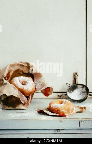 Hausgemachten Krapfen mit Zucker Pulver aus Paper Bag mit Vintage Sieb auf Blau Holztisch serviert. Rustikaler Stil, Tageslicht. Stockfoto