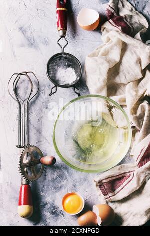 Zutaten und Utensilien zum Kochen von Baiser. Ungeschlacktes Eiweiß, Zuckerpulver, zerbrochene Eier, Vintage-Sieb und Handmixer auf Leinentuch über grau Stockfoto