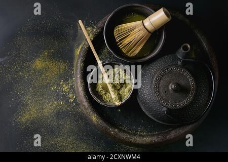 Grüner Tee Matcha-Pulver und Heißgetränk im schwarzen Schalen stehen mit Eisen-Teekanne, traditionelle Werkzeuge Bambuslöffel und Schneebesen in Terrakotta Schale über dunkel Stockfoto