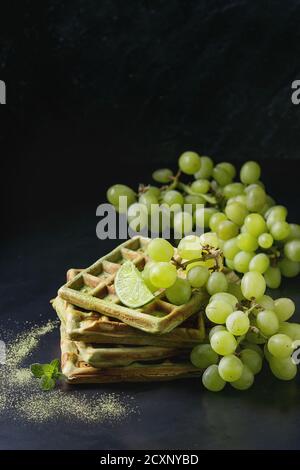 Hausgemachte quadratische grüne Matcha belgische Waffeln mit frischen reifen Trauben, Minze und Limette auf schwarzem Metall Hintergrund. Stockfoto