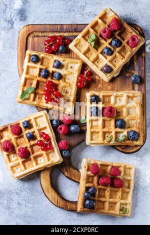 Hausgemachte quadratische belgische Waffeln mit frischen reifen Beeren Heidelbeere, Himbeere, rote Johannisbeere auf hölzernen Servierbrett über grauen Textur Hintergrund. Oben vi Stockfoto