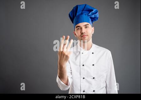 Porträt eines jungen Mannes in einem weißen Koch Anzug Zählen drei mit seinen Fingern posiert auf einem grauen Hintergrund gekleidet. Stockfoto