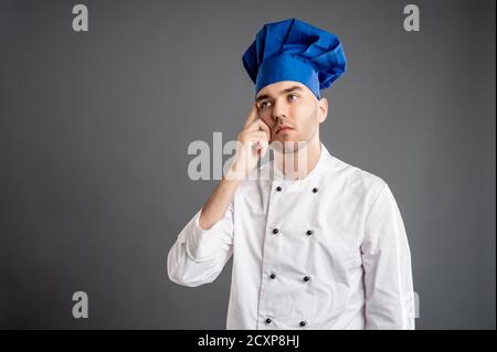 Porträt eines jungen Mannes in einem weißen Koch Anzug gekleidet Wundernd posiert auf einem grauen Hintergrund mit Kopie Raum Werbung Bereich Stockfoto