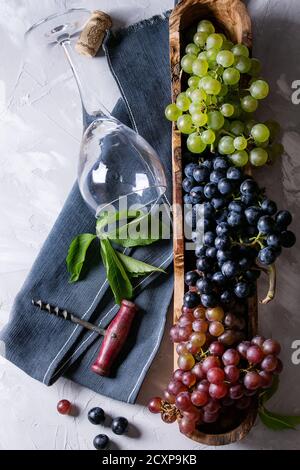 Vielzahl von drei Art frische reife Trauben dunkel Blau, Rot und Grün in Houten mit leeren Festlegung Wein Glas, alter Korkenzieher und grüne Blätter über Grau Stockfoto