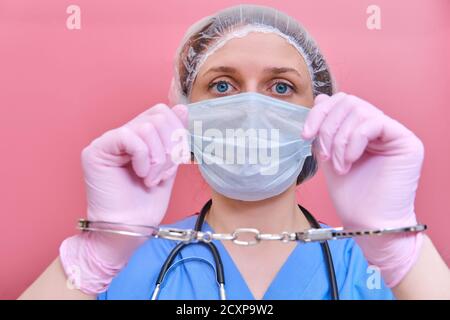 Porträt einer Ärztin in einer medizinischen Maske in Handschellen an den Händen, Nahaufnahme. Krankenschwester in blauer Uniform und Schutzhandschuhe auf rosa Hintergrund. Stockfoto