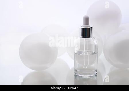 Kosmetisches Serum. Pipette mit einem Tropfen und einer Flasche und großen weißen Perlenkugeln auf einer weißen Spiegelfläche. Gesichtspflege. Stockfoto