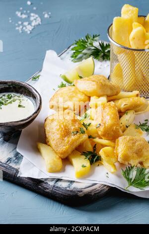 Traditionelle britische fast food Fisch und Chips. Mit weißem Käse Sauce, Kalk, Petersilie serviert, Pommes frites in Frittierkorbs auf weißem Papier über Blau co Stockfoto