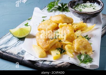 Traditionelle britische Fast-Food-Fisch und Chips. Serviert mit Weißkäse-Sauce, Limette, Petersilie, auf weißem Papier und schwarzem Servierbrett über blauem Beton Stockfoto