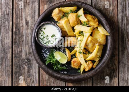 Traditionelle britische fast food Fisch und Chips. Mit weißem Käse Sauce, Kalk, Petersilie serviert, Pommes frites in den Frittierkorb in Terrakotta Fach über alte Stockfoto