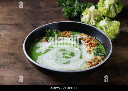 Vegetarische Brokkoli-Sahnesuppe in schwarzer Schüssel mit Sahne, gebratener Zwiebel, frischer Petersilie und Brokkoli auf altem Holzhintergrund serviert. Nahaufnahme. Gesund Stockfoto