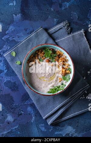 Pilzsahnesuppe in Keramikschale serviert mit Waldpilzen, Grünpflanzen, gebratene Zwiebel auf blauer Textilserviette über dunkelblauem Betonbackgroun Stockfoto