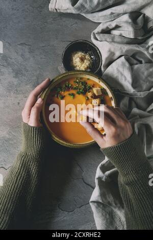 Schüssel vegetarische Kürbis Karotten Suppe serviert mit Safran Salz, Croutons und Zwiebeln auf Textil auf dunkelgrau Küchentisch. Weibliche Hände. Ansicht von oben mit der Stockfoto