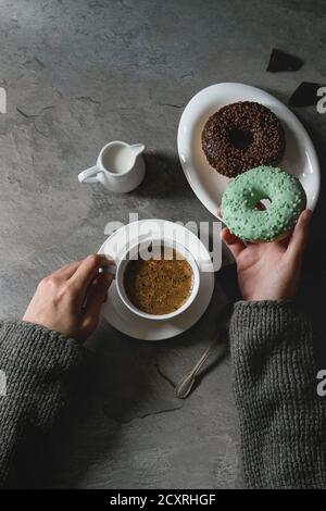 Teller mit bunten glasierten Donuts mit gehackter Schokolade, Tasse schwarzen Kaffee, Krug Milch über grauen Textur-Tisch. Weibliche Hände nehmen türkisfarbenen donat. Stockfoto