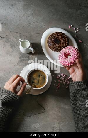 Platte von Bunt glasierter Krapfen mit rosa Zucker, Tasse schwarzen Kaffee, Kanne Milch über grau Textur Tabelle. Weibliche Hände Donat. Flach mit sp Stockfoto