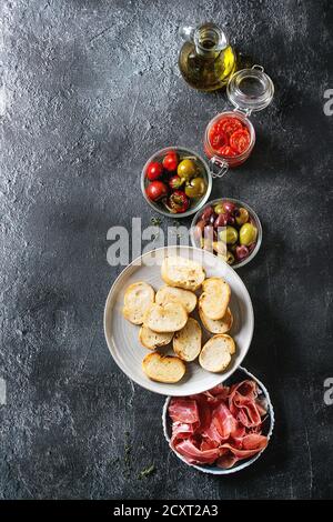 Zutaten für die Herstellung von Tapas oder Bruschetta. Knuspriges Brot, Schinken, Prosciutto, sonnengetrocknete Tomaten, Olivenöl, Oliven, Paprika, Grüns auf Platten über dunkle te Stockfoto