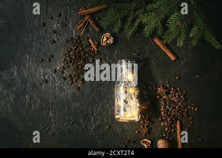 Weihnachtsbeleuchtung Girlande in Glasflasche mit Kaffeebohnen, Gewürze, Nüsse, cocktail Rohr, Fir Tree Branches in Holz- fach über dunkle Textur Hintergrund Stockfoto