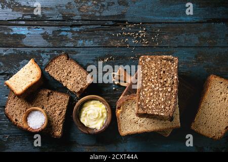 Verschiedene Brote in Scheiben hausgemachtes Roggenbrot Vollkorn und Samen für das Frühstück mit Olivenholz Schüsseln mit Butter und Salz über Altes, dunkles Holz- backgrou Stockfoto