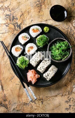 Hausgemachte Sushi-Brötchen mit Lachs, Sesam serviert in schwarzem Teller mit rosafarbenem Ingwer, Sojasauce, Wasabi, Algensalat, Essstäbchen über Stockfoto