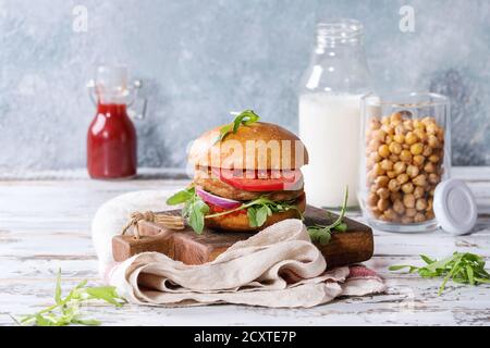 Hausgemachten Burger im klassischen Brötchen mit Tomaten, Rucola, Fleisch, Käse, Zwiebel auf Holzbrett mit einer Flasche Ketchup, Milch serviert, geröstete Kichererbsen ove Stockfoto