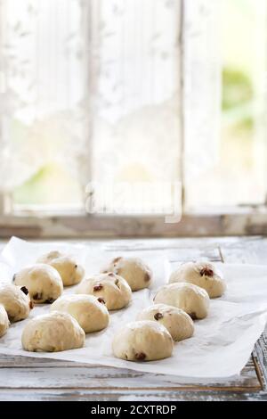 Raw ungebackene Gebäck. Bereit, hausgemachte Ostern traditionelle Hot Cross Buns auf Backpapier über weiße Holztisch mit Fenster im Hintergrund zu backen. Natürliche Stockfoto