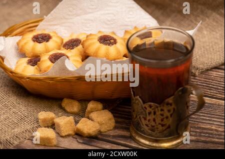 Hausgemachte Gebäck-Kekse mit Marmelade und einem facettierten Glas Tee in einem Vintage-Cup-Halter auf einem Hintergrund von homespun Stoff mit einer rauen Textur, close-up, Stockfoto