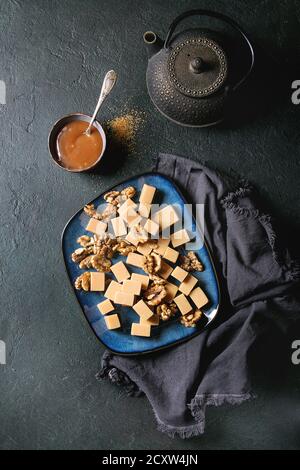 Gesalzene caramel fudge Candy auf blaue Platte mit Karamell Sauce serviert, karamellisierte Walnüsse, textile Serviette und Eisen Teekanne über schwarzen Textur Hintergrund. Stockfoto