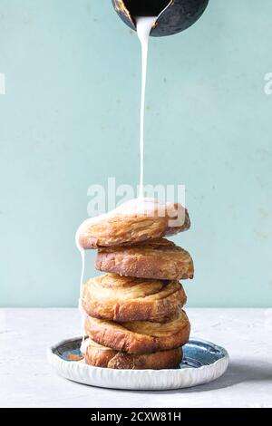 Stapel von hausgemachten verglaste Blätterteig Zimtschnecken mit Vanillesauce und Rosinen auf blauen Platte über Grau grün Hintergrund. Gießen Glasur aus der Kanne. Pin-up Stockfoto