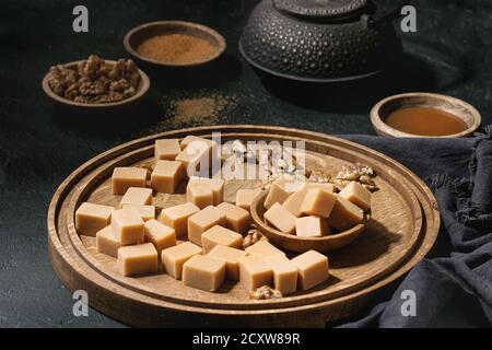 Gesalzene caramel fudge Candy auf Holzbrett mit Karamell Sauce serviert, braunen Rohrzucker und karamellisierte Walnüsse in Holz Schalen, Bügeleisen Teekanne über Schwarzes t Stockfoto