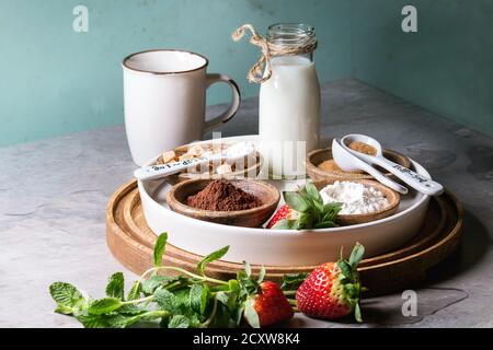 Zutaten zum Kochen Schokolade Tasse Kuchen. Mehl, Kakao, Zucker, Karamell in Holzschalen, Milch, Erdbeeren und Minze auf rundes Tablett Esprit serviert. Stockfoto