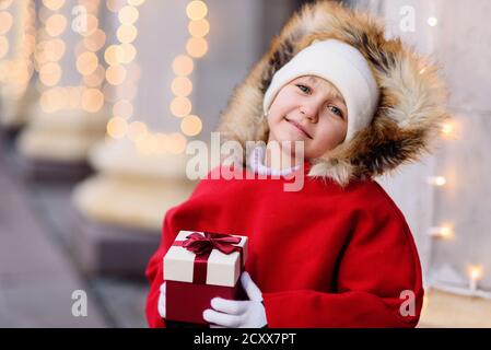 Kind auf weihnachtsmarkt. Nettes schönes Mädchen in roten Hut auf der Straße glücklich über Geschenk-Box stehen. Traditionelle Freizeit für Familien an Weihnachten. Stockfoto