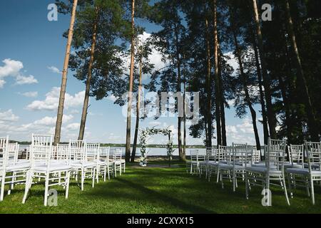 Hochzeitszeremonie im Freien an der Seeufer - Hochzeit Bogen mit frischen Blumen und leeren weißen Stühlen dekoriert Stockfoto