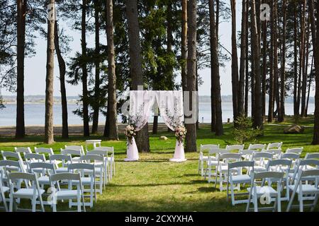 Ein mit frischen Blumen und leeren Stühlen geschmückter Hochzeitbogen, der für eine Hochzeit im Freien im Wald nahe der Seeufer vorbereitet ist. Stockfoto