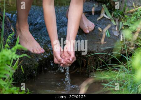 Kleiner Junge, der an einem kleinen Bach mit klarem Wasser spielt Mit seinen Händen und Füßen mit der Wasserkühlung Und erfrischend mit reinem Elixier des Lebens Stockfoto