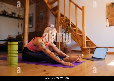 Junge Frau mit einer Tätowierung und Schaumstoffrolle zur Seite. Fitness virtuelle Online-Übung. Stretching aus dem Internet zu Hause. Ein sportliches Mädchen in Sportswear Stockfoto