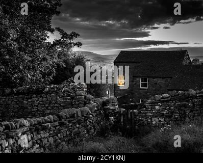 Schwarz-Weiß-Bild eines Steinhauses in der Abenddämmerung mit warmen orangefarbenen Leuchten aus einem Fenster in den Yorkshire Dales, England, Großbritannien. Stockfoto