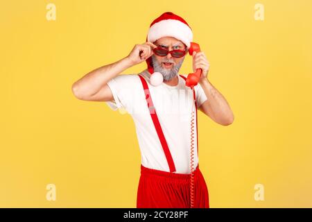 Brutal grau bärtig erwachsenen Mann in weihnachtsmann Hut hält Hörer des Festnetztelefons Blick auf die Kamera durch Sonnenbrille, rufen zu gratulieren. Stockfoto