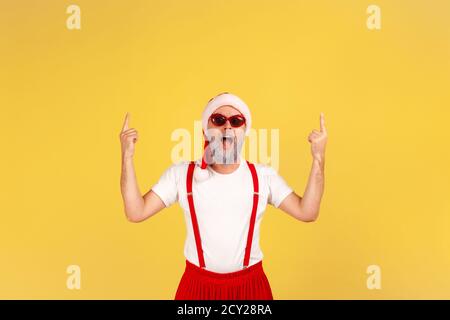 Extrem glücklich positive ältere Mann in weihnachtsmann Hut und stilvolle Sonnenbrille zeigen Finger nach oben auf leere Stelle für Ihre Werbung. Studio im Innenbereich Stockfoto