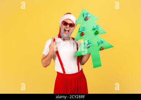 Extrem glücklich aufgeregt grauen bärtigen Mann in weihnachtsmann Hut hält Papier weihnachtsbaum zeigt Rock and Roll Geste, Spaß an den Winterferien. Stockfoto