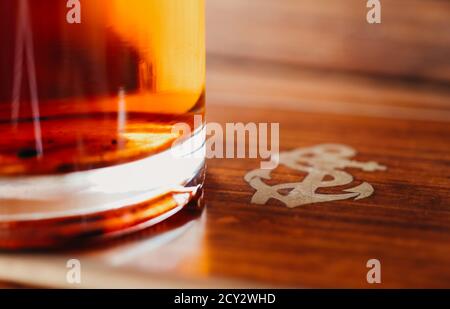 Fragment eines Glases mit altem Rum auf einem hölzernen Oberfläche mit dem Bild eines Seeankers aus nächster Nähe Mit flachem Freiheitsgrad Stockfoto