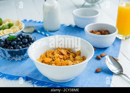 Nahrhaftes, gesundes Frühstück. Schüssel mit Getreide, Mandeln, Heidelbeere, Milch auf pflanzlicher Basis und Orangensaft auf blauer Serviette und weißem Holztisch Stockfoto